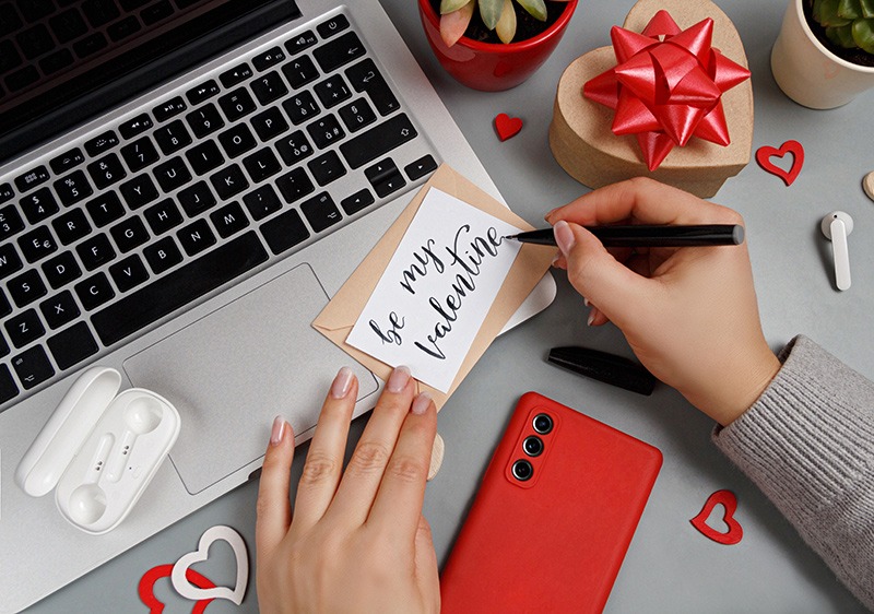 woman writing valentines day card near laptop and smart phone